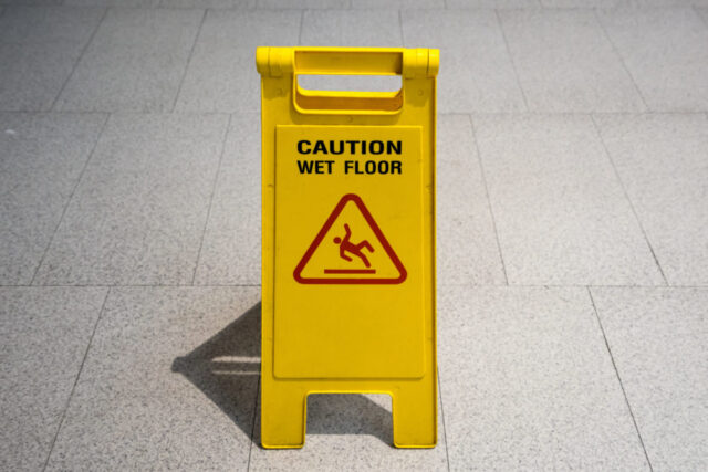 A yellow "Caution Wet Floor" sign stands prominently on the tiled floor, featuring a warning symbol of a person slipping inside a red triangle, alerting passersby to potential premises liability accidents.