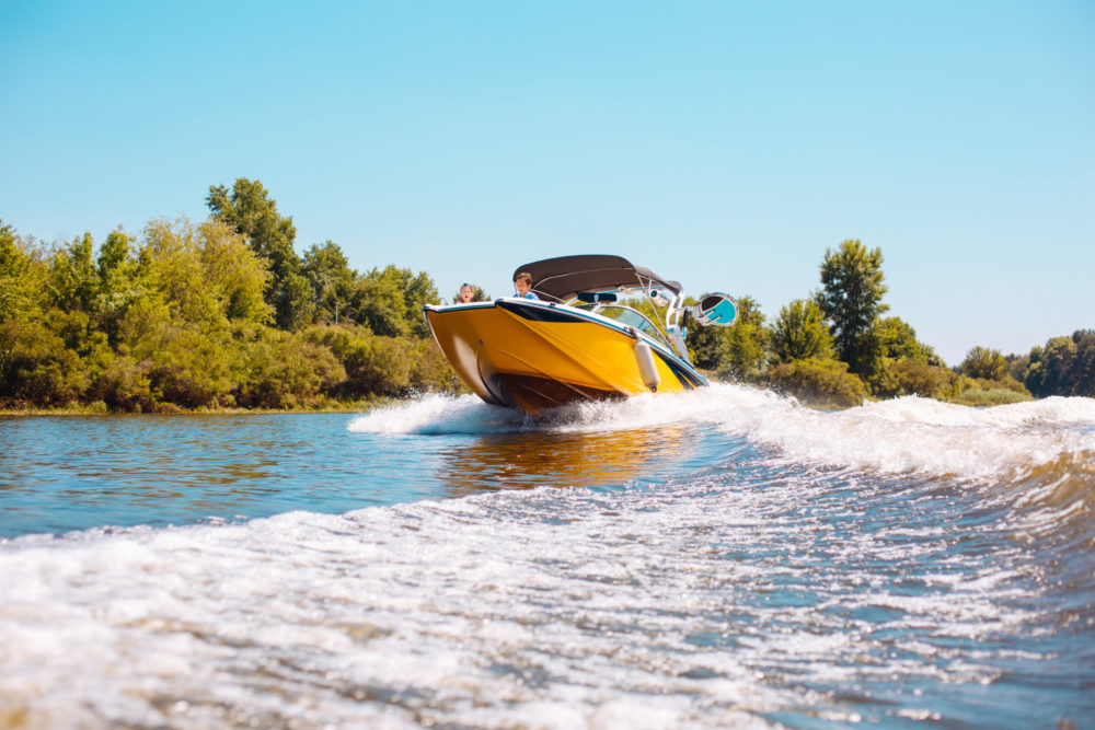 A yellow motorboat speeds along a river, creating a trail of white waves as it weaves through the water. Surrounded by green trees under a clear blue sky, it paints a picturesque scene—a world where boating accidents are far from the mind.
