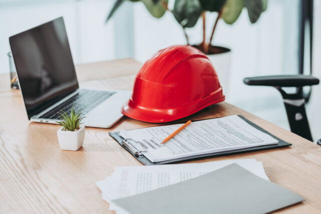 A desk with a laptop, a red safety helmet—a reminder of workplace accidents—documents on a clipboard with a pencil, and a small potted plant. A chair is partially visible in the background, illuminated by natural light from a nearby window.