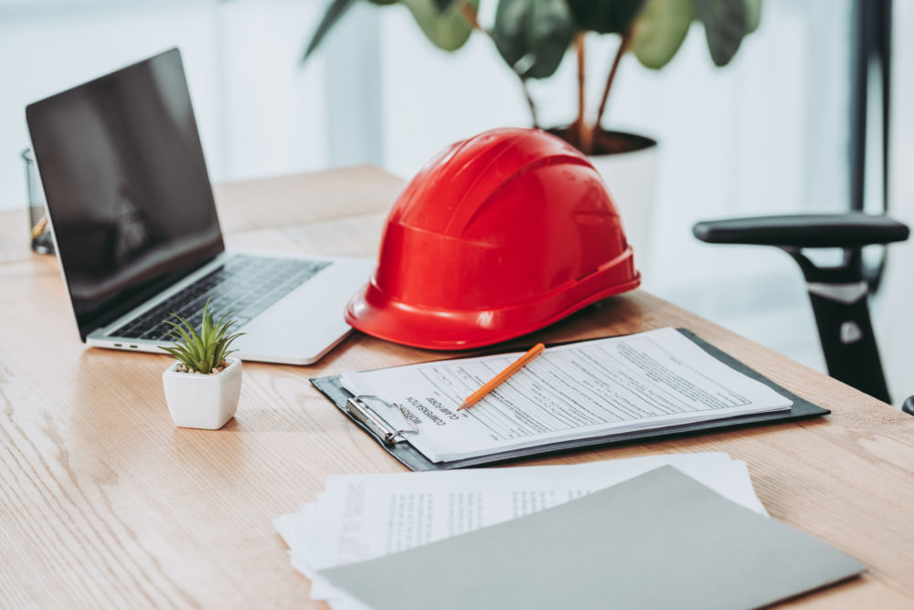 A desk with a laptop, a red safety helmet—a reminder of workplace accidents—documents on a clipboard with a pencil, and a small potted plant. A chair is partially visible in the background, illuminated by natural light from a nearby window.