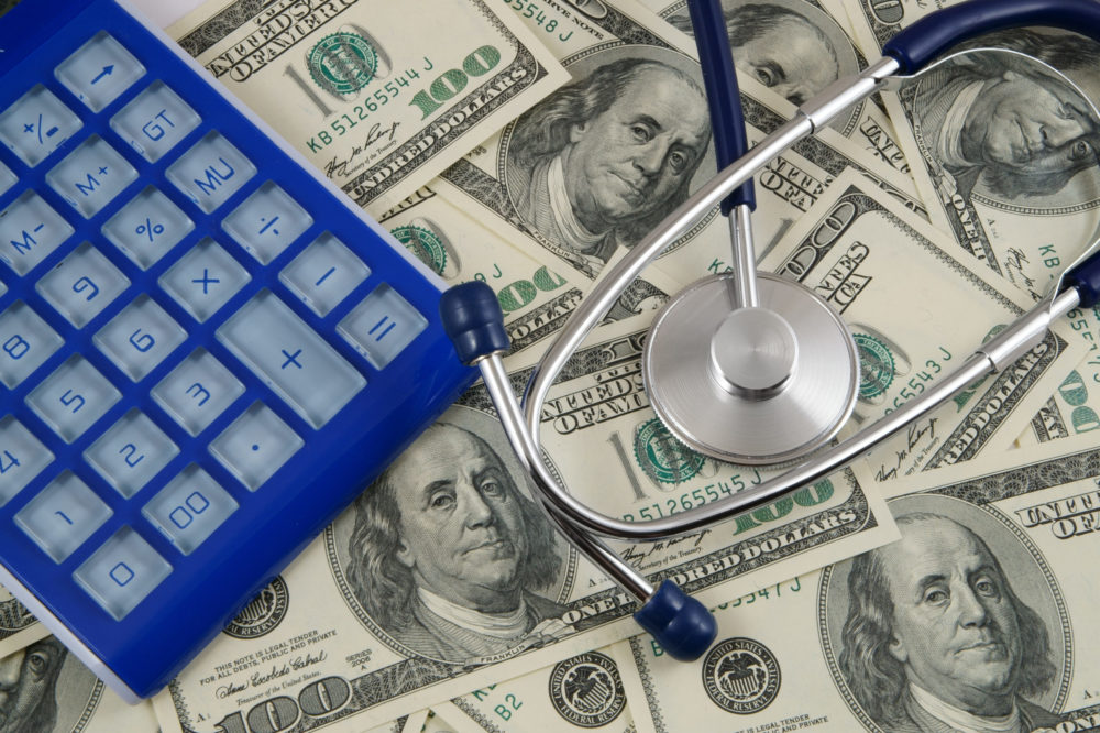 A blue calculator and a stethoscope rest on a pile of one hundred dollar bills, symbolizing the potential loss of earnings in the healthcare sector.