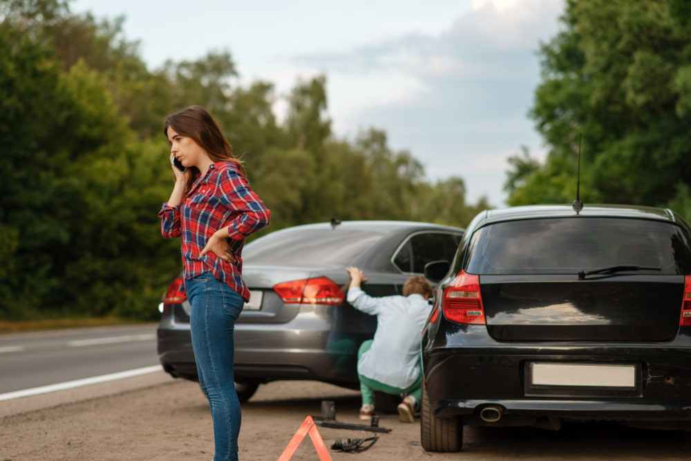 A woman in a plaid shirt stands by the roadside, talking on her phone, likely concerned about an uninsured driver. In the background, a person crouches to change the tire of a dark-colored car. Nearby are an emergency warning triangle and a car jack. Trees line the road.