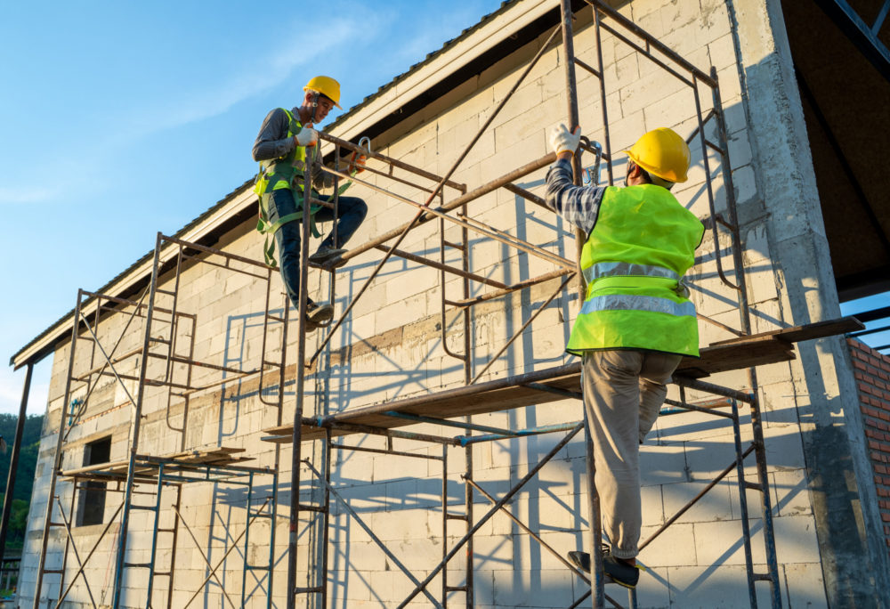 Two construction workers in safety gear and yellow helmets are diligently working on scaffolding beside a partially built structure, mindful of potential scaffolding accidents. One worker is climbing while the other securely holds the structure, with sunlight illuminating the scene.