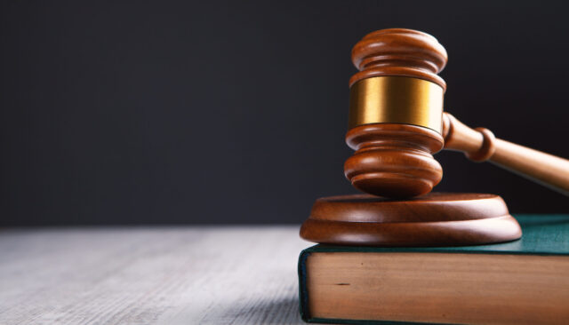 A wooden judge's gavel rests on a thick, closed book against a dark background, ready to hammer down justice. Positioned slightly to the right with its handle extending off the book's surface, it conveys an immediate sense of legal proceedings, as if ready to address an urgent report.