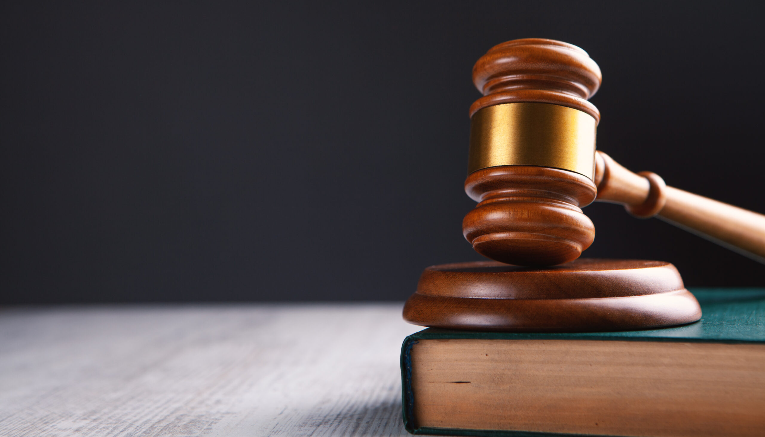 A wooden judge's gavel rests on a thick, closed book against a dark background, ready to hammer down justice. Positioned slightly to the right with its handle extending off the book's surface, it conveys an immediate sense of legal proceedings, as if ready to address an urgent report.