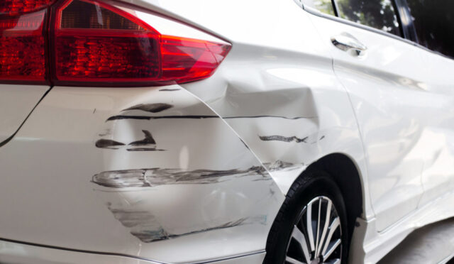 A white car shows signs of a hit and run, with dents and black scrape marks on the rear left side. Despite the damage, the taillight remains intact, and a wheel with a black and silver rim is partially visible.