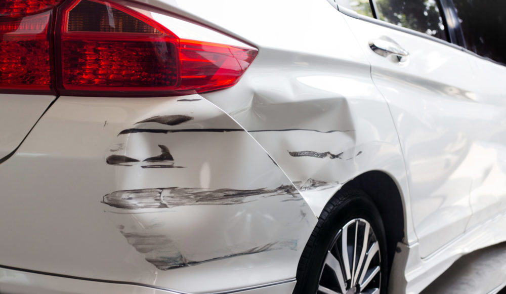 A white car shows signs of a hit and run, with dents and black scrape marks on the rear left side. Despite the damage, the taillight remains intact, and a wheel with a black and silver rim is partially visible.