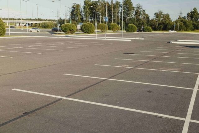 An empty parking lot with clearly marked parking spaces. Several cars are visible in the distance, surrounded by grassy areas and trees. The sky is clear.