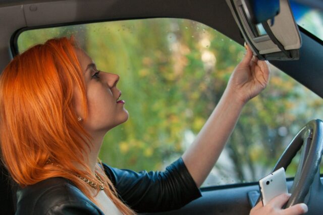 A person with long red hair sits in a car, adjusting the rearview mirror. They hold a smartphone in their other hand. The background shows a blurred view of trees.