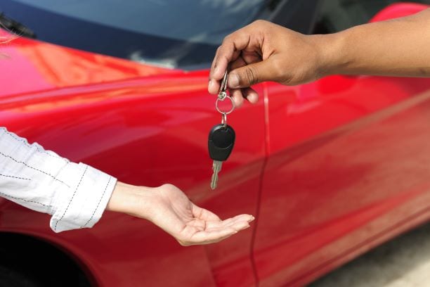 A close-up image of a person handing car keys to another person. The red car is visible in the background. The person receiving the keys is wearing a white shirt with stripes.