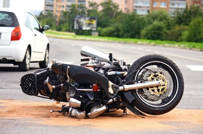A motorcycle lies on its side on a road, surrounded by sand, possibly to absorb fluids. A white car is parked nearby, and trees and buildings are visible in the background.