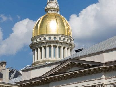 The image shows a domed building with a golden top and white columns, set against a blue sky with clouds. The architectural style is classical, featuring a prominent rotunda.