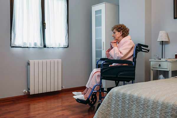 An elderly person in a wheelchair wearing a pink robe sits in a bedroom, gazing out of a window. The room has wooden floors, a radiator, and a cabinet. A bedside table with a lamp is next to the bed.