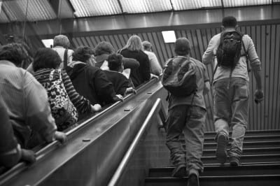 A group of people, some with backpacks, ascends a staircase in a public building. The photo is black and white, capturing the scene from behind as they move upward, with a slatted ceiling above them.