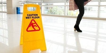 A yellow caution sign indicating a wet floor is placed on a glossy, light-colored tiled surface. In the background, a person wearing black boots and checkered clothing is walking past the sign. Large windows allow natural light into the area.