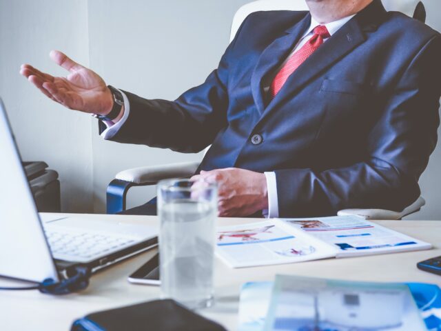 A personal injury attorney in a business suit sits at a desk with a laptop, smartphone, and open documents during an initial consultation. A glass of water is on the desk. The attorney is gesturing with their right hand while facing slightly away from the camera, perhaps in an office in New Jersey.