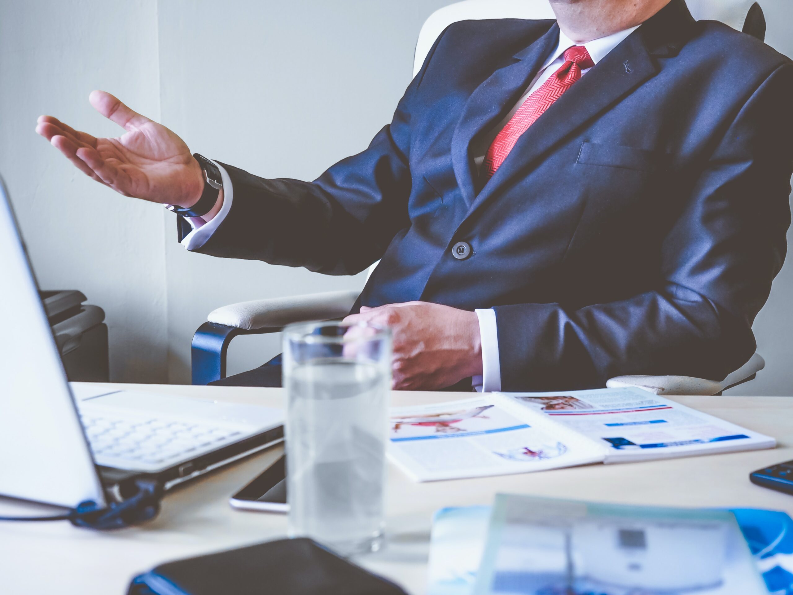 A personal injury attorney in a business suit sits at a desk with a laptop, smartphone, and open documents during an initial consultation. A glass of water is on the desk. The attorney is gesturing with their right hand while facing slightly away from the camera, perhaps in an office in New Jersey.