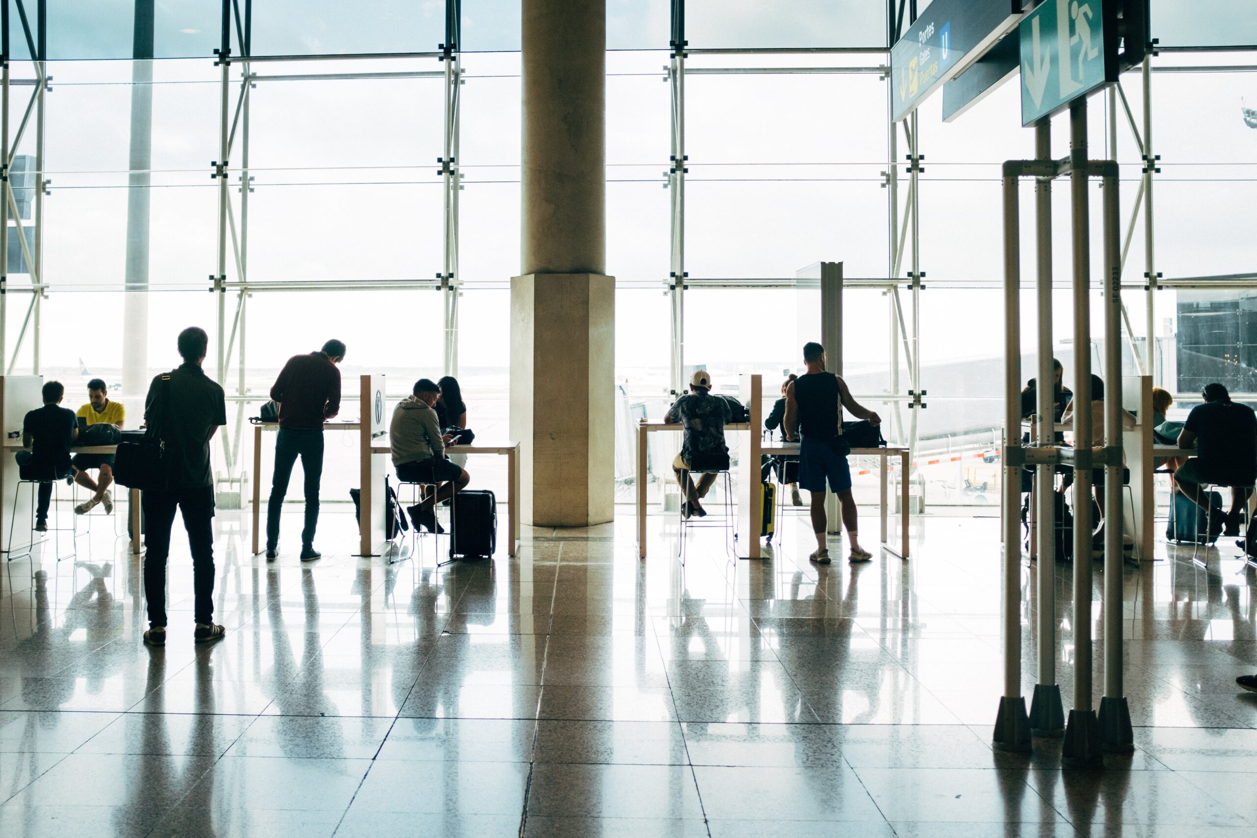 People are sitting and standing near a large window in an airport terminal. Some are using electronic devices, and others are at tables. The bright natural light streams through the windows, casting reflections on the shiny floor—a scene far removed from thoughts of airport accident attorneys.