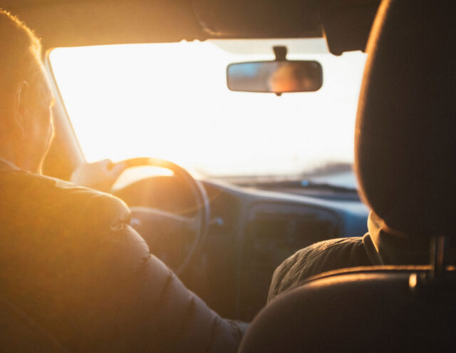 A person is driving a car during sunset. The sun's rays create a warm glow inside the vehicle, illuminating the driver's left shoulder. The dashboard and part of the passenger seat are visible in the foreground.
