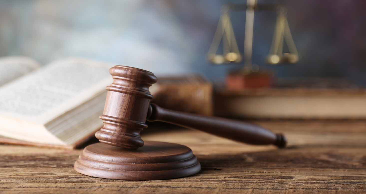 A wooden gavel rests on its base on a wooden table. Behind it, an open book and a blurred set of scales are visible. The image suggests a legal or judicial theme.