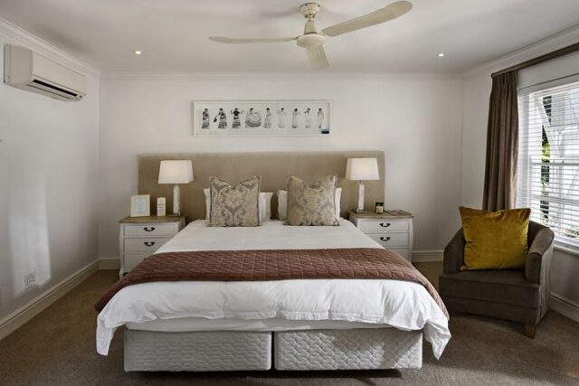 A cozy Airbnb bedroom showcases a large bed with white linens and a brown throw, complemented by two pillows against a beige headboard. Two side tables with lamps flank the bed. By the window, blinds filter the light over a brown armchair with a yellow cushion, while a ceiling fan whirls above.