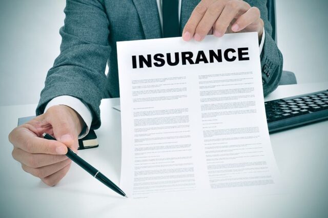 A person in a suit holds an insurance document and a pen over a desk. A keyboard and a notebook are partially visible.