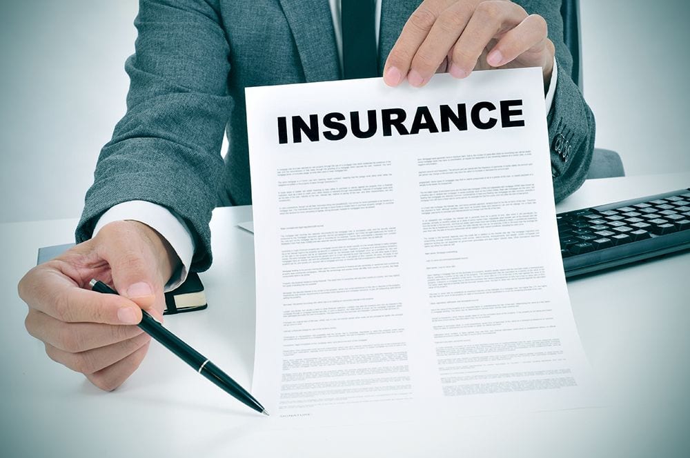 A person in a suit holds an insurance document and a pen over a desk. A keyboard and a notebook are partially visible.