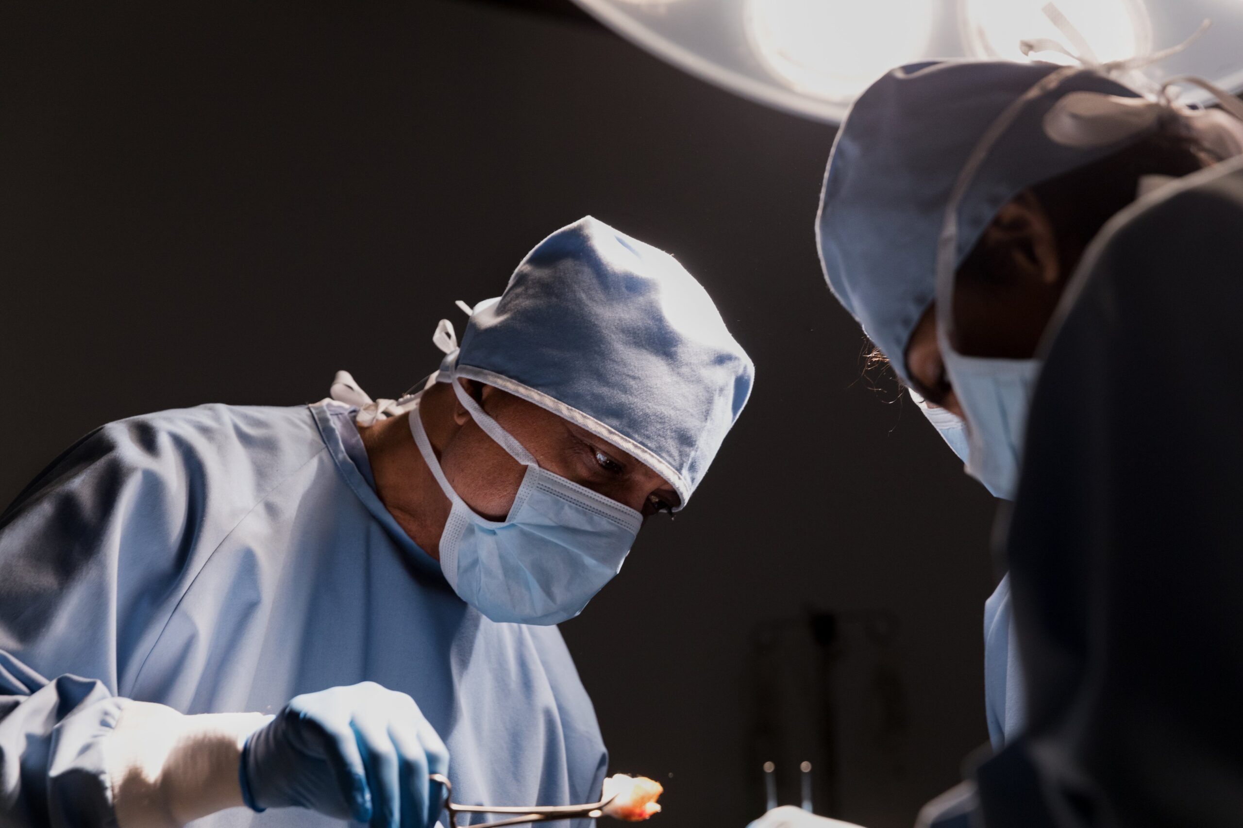 Two surgeons in blue scrubs and masks perform a surgical procedure under bright lights. One holds surgical tools, focusing intently on their task—a scene reminiscent of the precision needed by brain injury attorneys. The dimly lit background emphasizes the operating room setting.