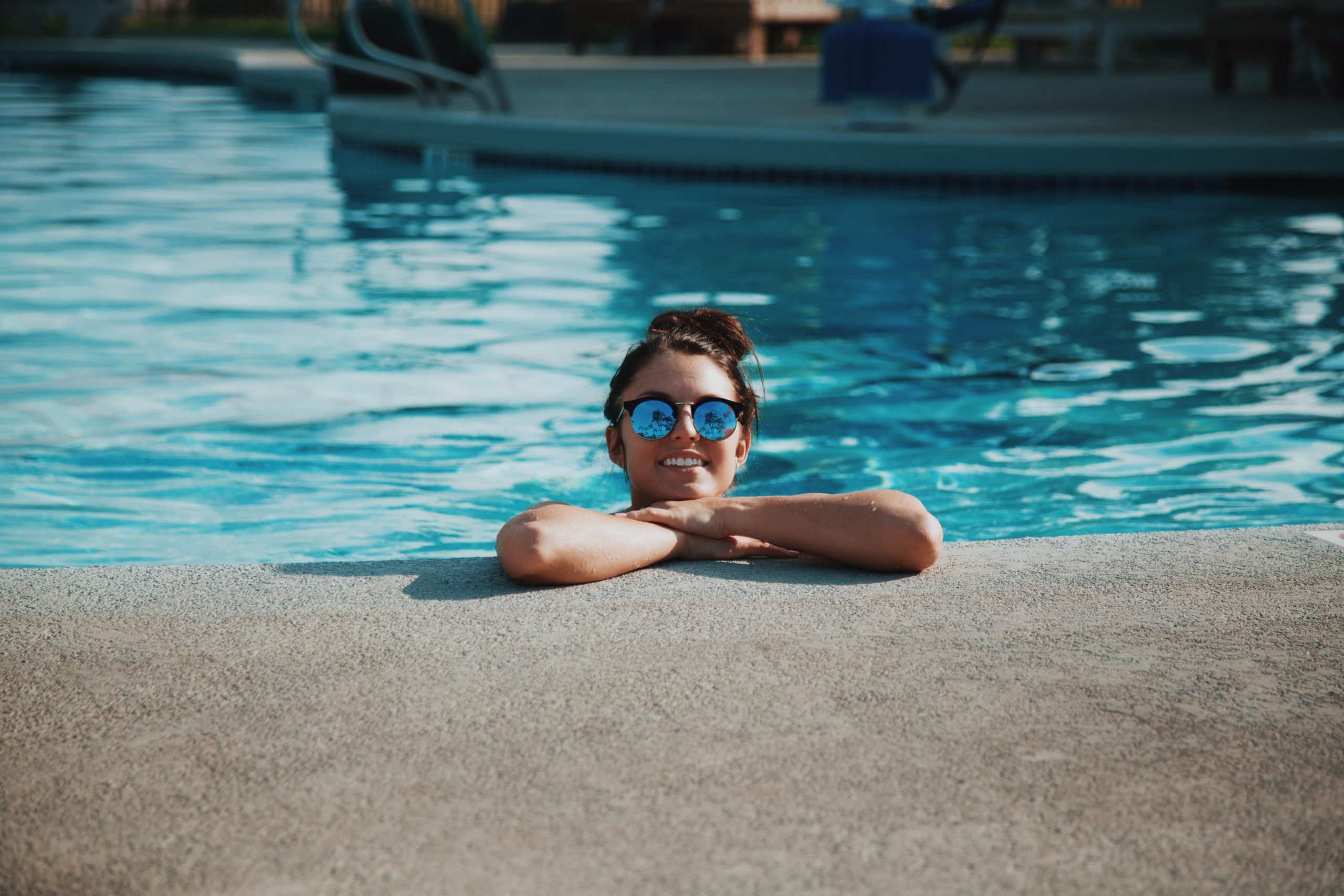 A person with dark hair wearing reflective sunglasses is partially submerged in a swimming pool, resting their arms on the edge. Despite the sunny day inviting relaxation, they're mindful of pool accidents while enjoying the outdoors.