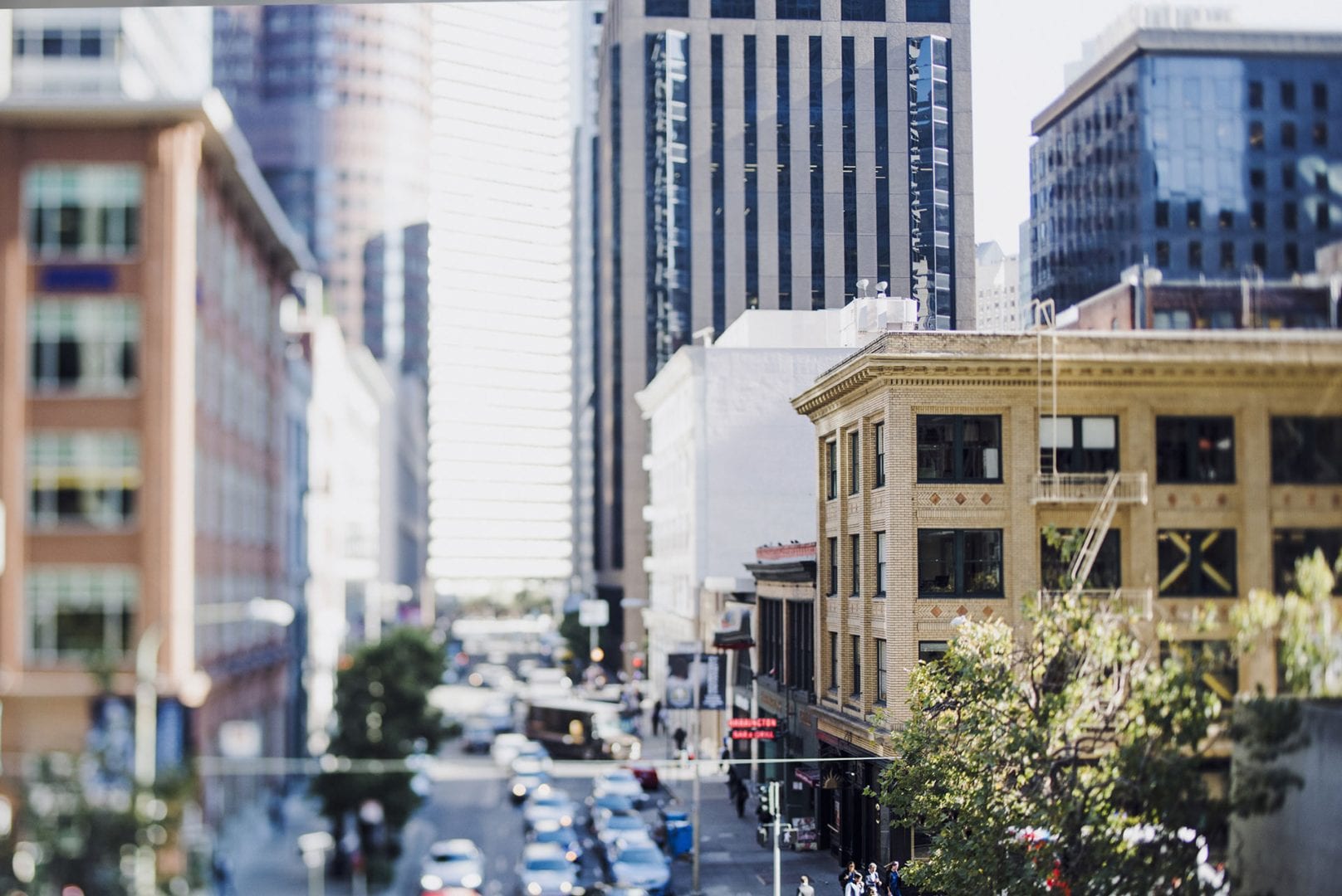 A busy city street is lined with multi-story buildings under a clear sky. The scene includes cars, pedestrians, and greenery, with sunlight illuminating the facades of the buildings.