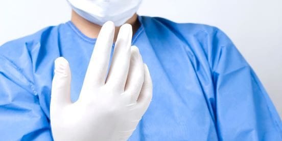 A person wearing a blue surgical gown and a mask is holding up a hand covered with a white latex glove, set against a plain white background.
