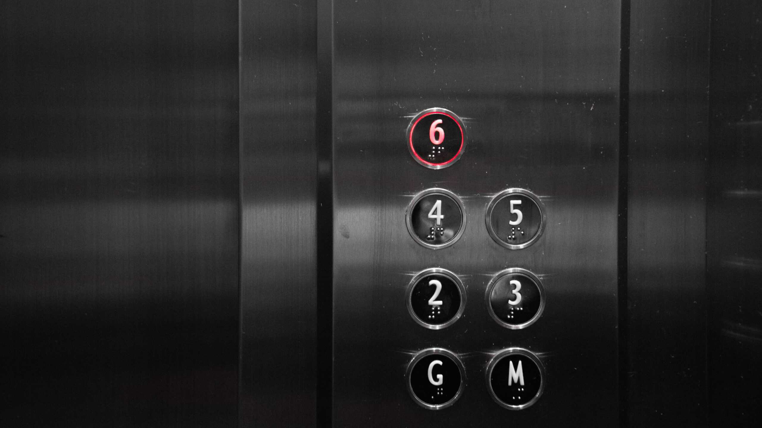 Elevator button panel with floors labeled G, M, 2, 3, 4, 5, and 6. The button for floor 6 is lit in red, indicating selection. The metallic panel features braille under each number. A reminder of the importance of safety standards often highlighted by elevator accident attorneys.