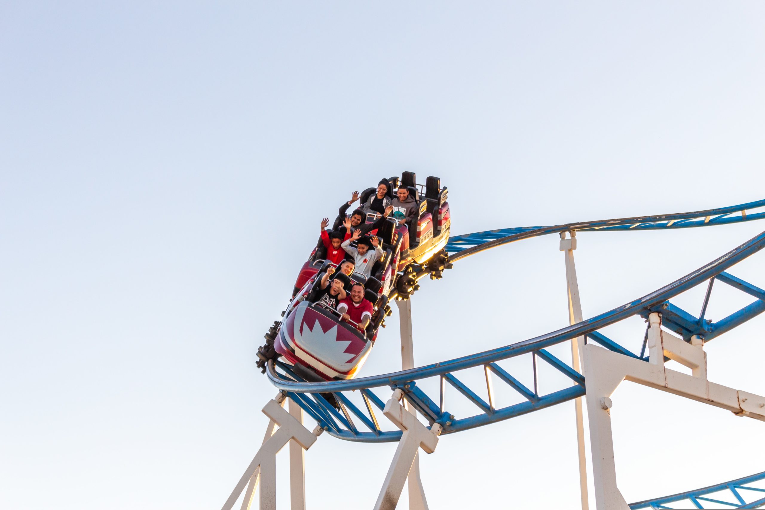 A roller coaster, untouched by amusement park accidents, ascends on blue and white tracks against a clear sky as people enjoy the ride. Seated in a red car, the riders' excitement is infectious as they raise their hands high.