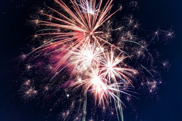 A vibrant fireworks display featuring red, white, and pink bursts illuminates the night sky on the Fourth of July, creating a dazzling spectacle with sparkling trails and colorful explosions.