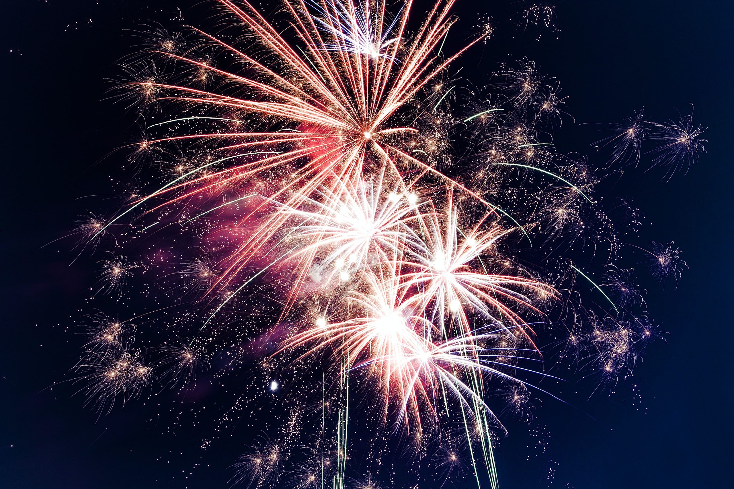 A vibrant fireworks display featuring red, white, and pink bursts illuminates the night sky on the Fourth of July, creating a dazzling spectacle with sparkling trails and colorful explosions.