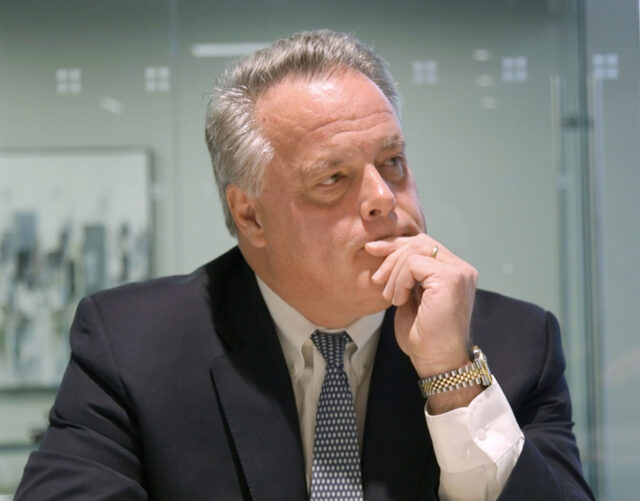 A man with gray hair is sitting at a desk, wearing a dark suit, white shirt, and patterned tie. He has his hand on his chin, appearing thoughtful. There is a blurred painting and glass wall in the background.