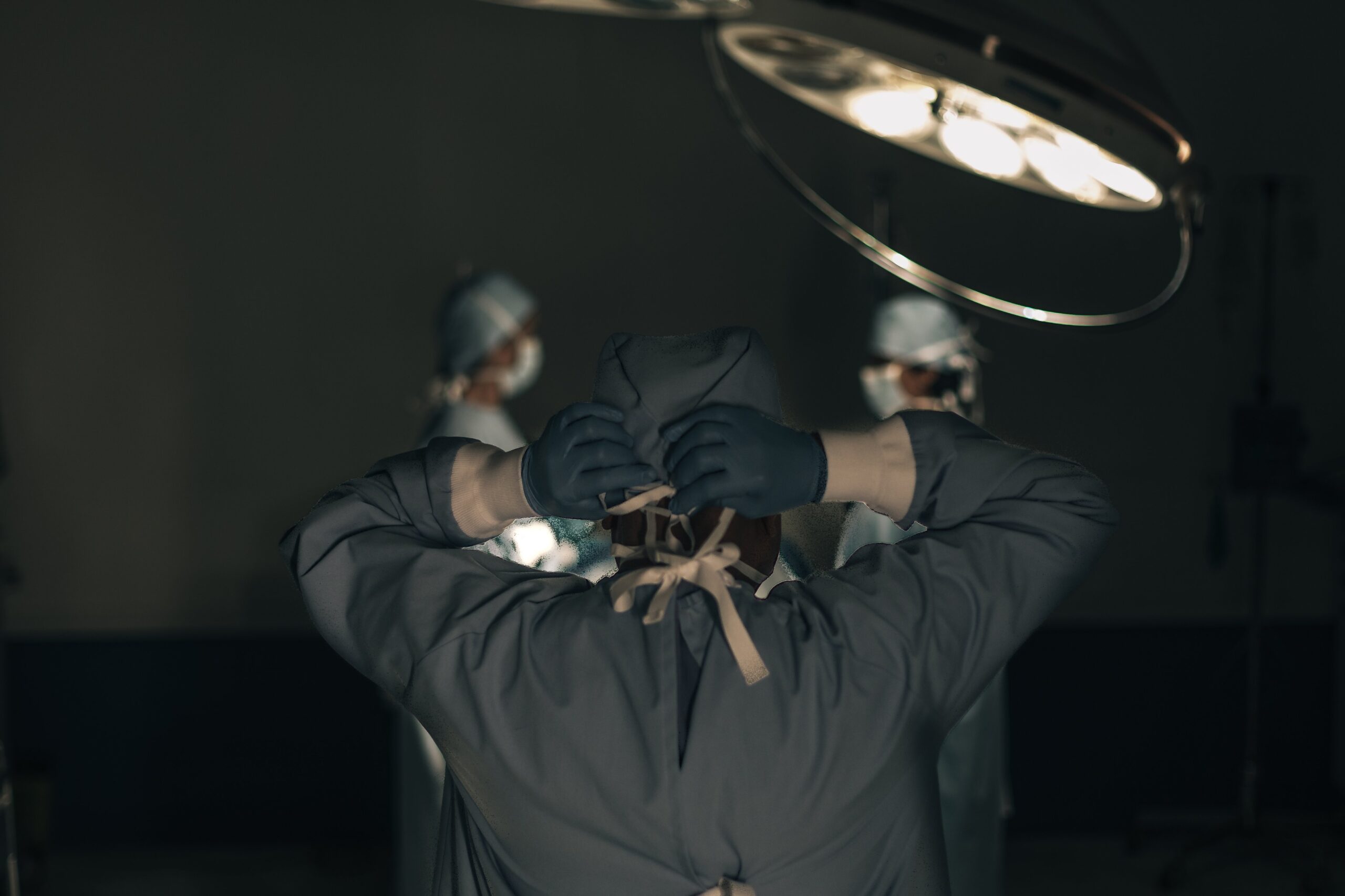 A surgeon, seen from behind, adjusts their cap under an overhead surgical light. Two other medical professionals, also in scrubs, are in the background, focused on a procedure in what appears to be an operating room—a scene that underscores the importance of precision for patients and medical malpractice attorneys alike.