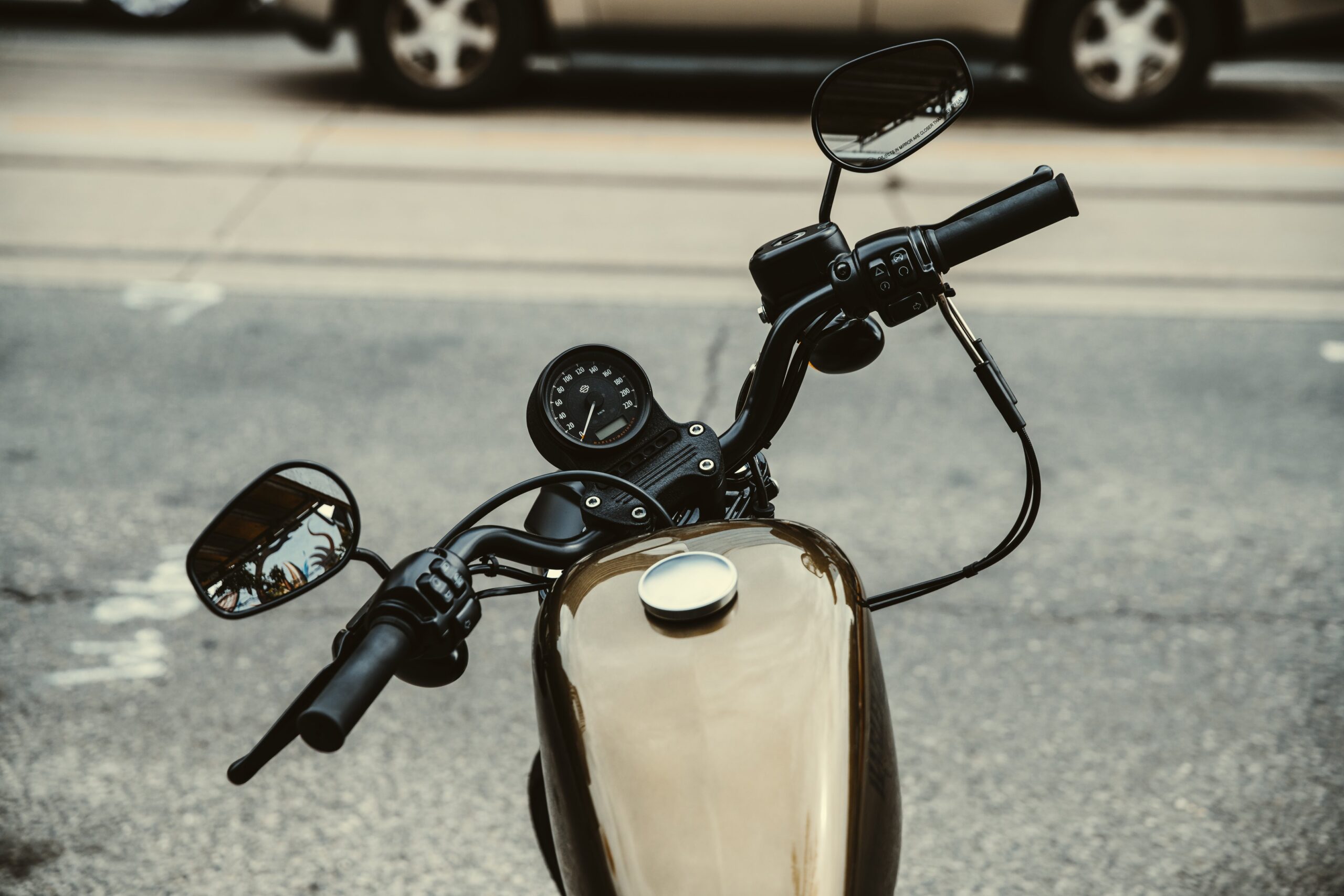 A close-up view of a motorcycle's handlebars and dashboard, showcasing rear-view mirrors and the speedometer. Parked on a street with blurred cars in the background, it subtly highlights the importance of safety—a reminder that motorcycle accident attorneys are essential for riders. The fuel tank gleams in the foreground.