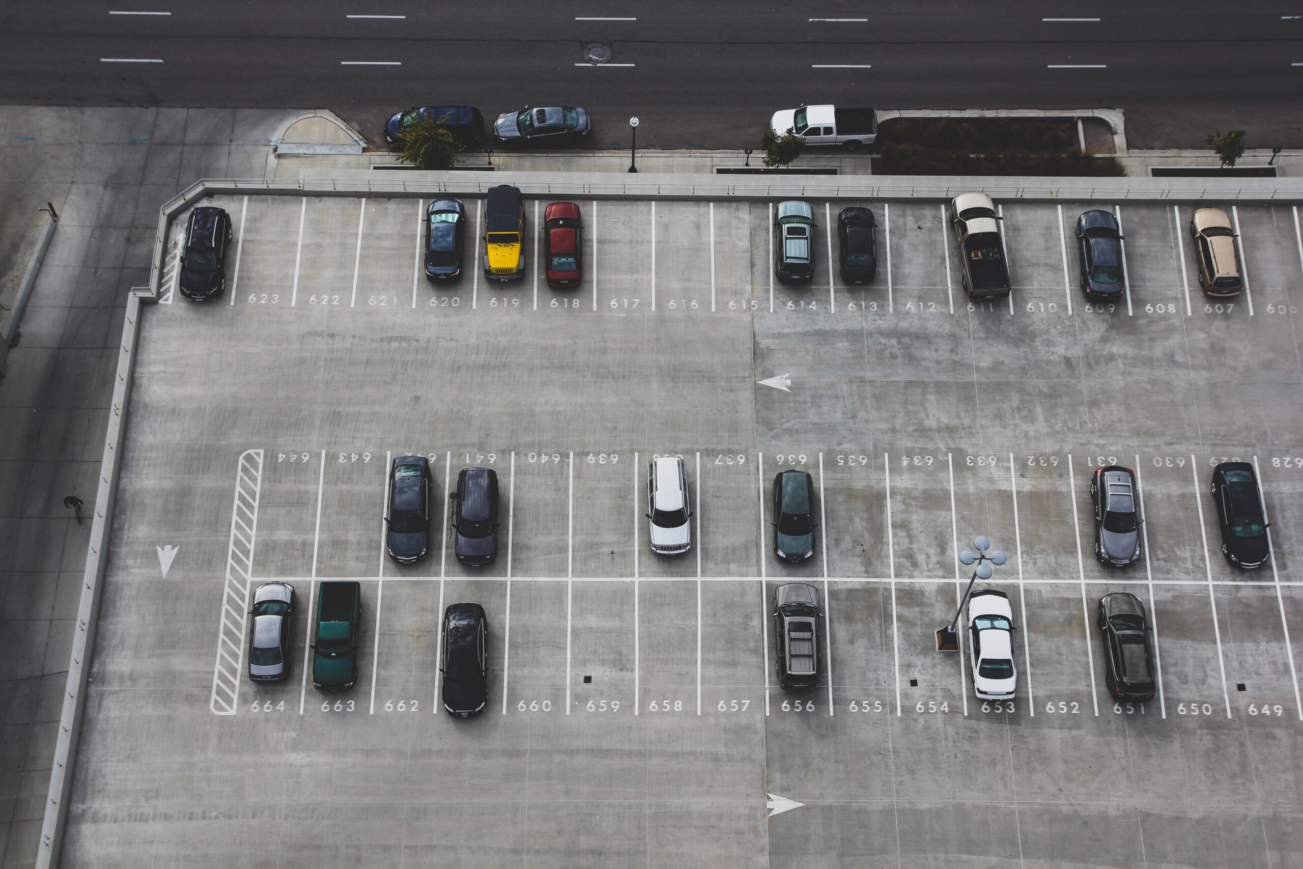 An overhead view shows a parking lot with two rows of parked vehicles on a concrete surface, meticulously marked by white lines and numbers designating each space. A road with several lanes runs parallel to the lot at the top of the image, where, thankfully, no parking lot accident attorneys are needed today.