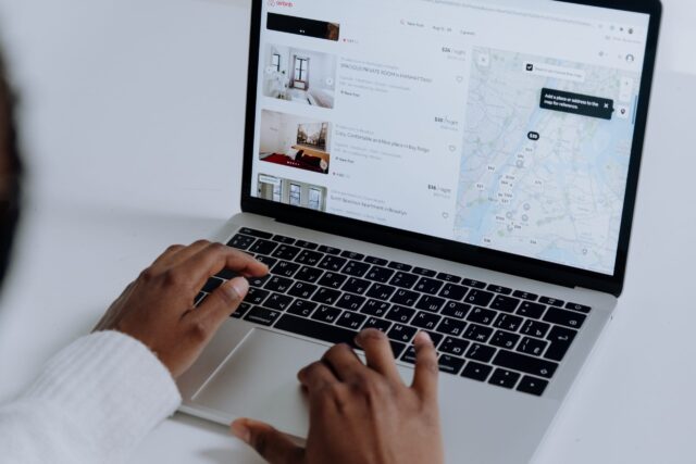 A person is using a laptop to browse a rental listing website, much like Airbnb. The screen displays various property options with prices and a map highlighting locations. Focused and intent, the person types diligently on the keyboard, carefully reviewing each option to avoid any potential mishaps.
