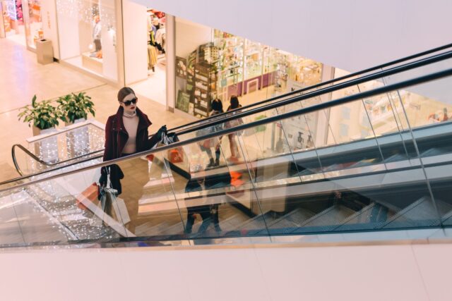 In a bustling New Jersey shopping mall, a person wearing sunglasses and a red coat ascends an escalator, holding a shopping bag. The background features vibrant store displays and decorative lights, creating the perfect shopping atmosphere.