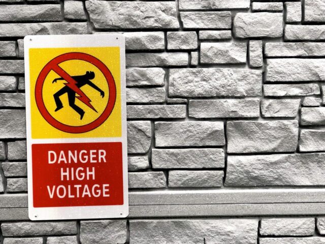 A high voltage warning sign on the stone wall shows a silhouette enduring electrocution from a lightning bolt, with "Danger High Voltage" written below.