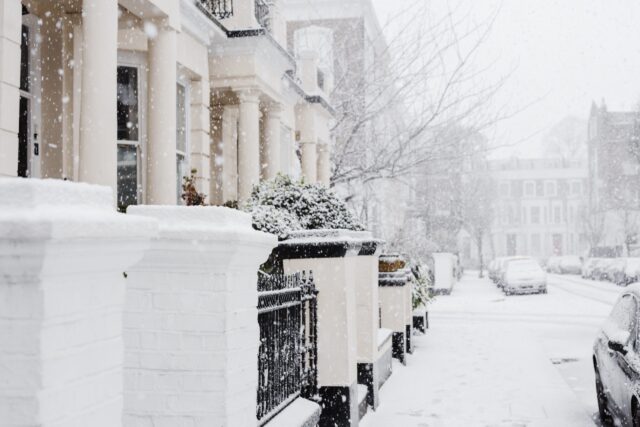A row of residential houses with columns is seen during a snowfall. Snow covers the ground, trees, and cars parked along the street, offering a chilly reminder of snow and ice removal duties. The scene looks cold and wintry.