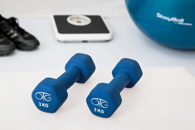 The image depicts two blue 2 kg dumbbells on a white surface. In the background, an exercise ball, a pair of black sneakers, and a scale suggest a fitness setting. The scene subtly hints at potential injuries from defective home gym equipment if not used with care.
