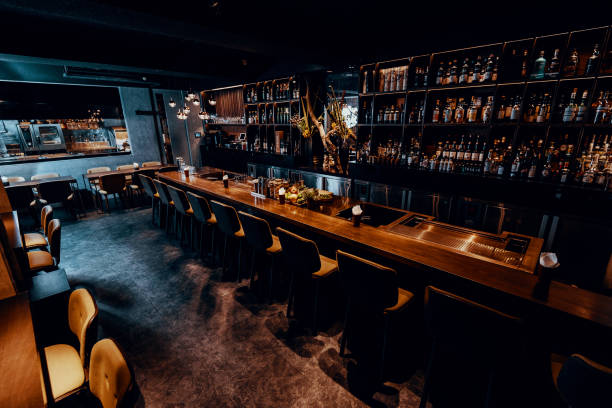 A bar with poor lighting features a long wooden counter lined with high chairs. Behind the bar, shelves display various bottles. The seating area includes tables and chairs, creating a warm and inviting ambiance despite the dimly lit surroundings.