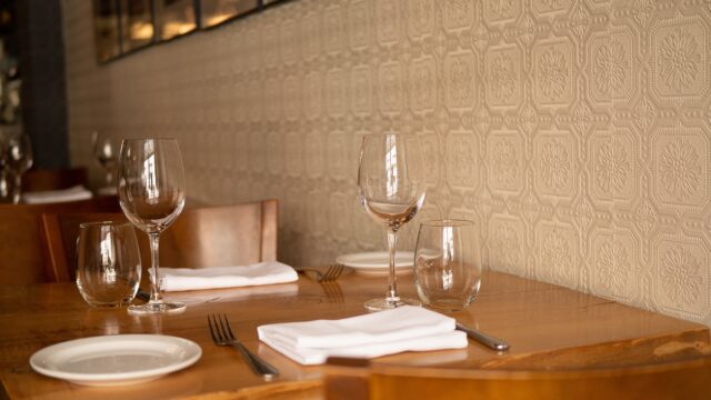 A restaurant table set for two with wooden chairs includes empty wine glasses, water glasses, white plates, and cutlery with neatly folded napkins. The wall's textured geometric pattern adds charm to the intimate setting. For any unforeseen mishaps, remember Restaurant Accident Attorneys are just a call away.