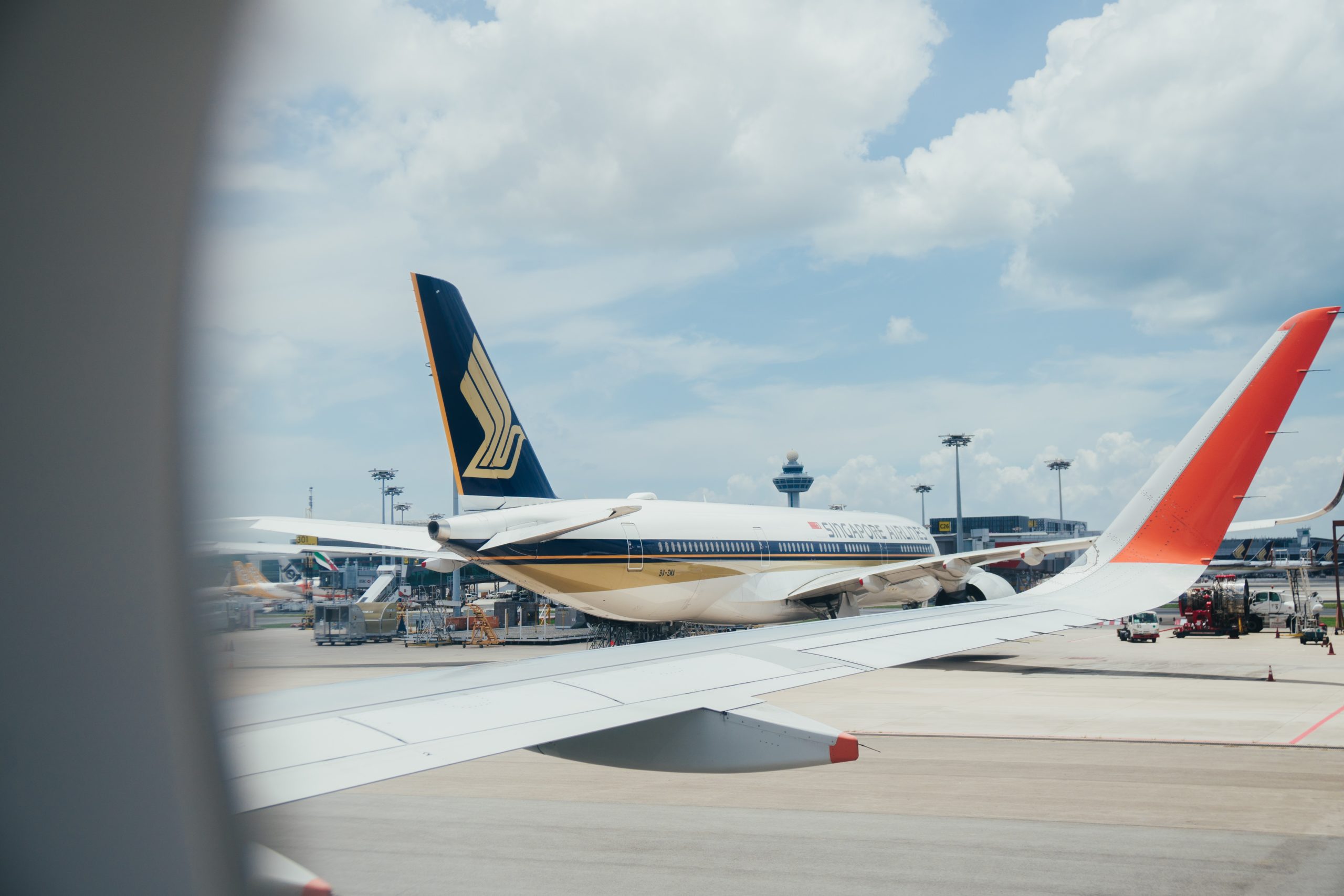 View from an airplane window showing a Singapore Airlines aircraft parked at the airport gate, with its distinct tail design visible. The airport control tower and other planes are in the background under a partly cloudy sky, reminding travelers to stay mindful of potential airplane injuries.