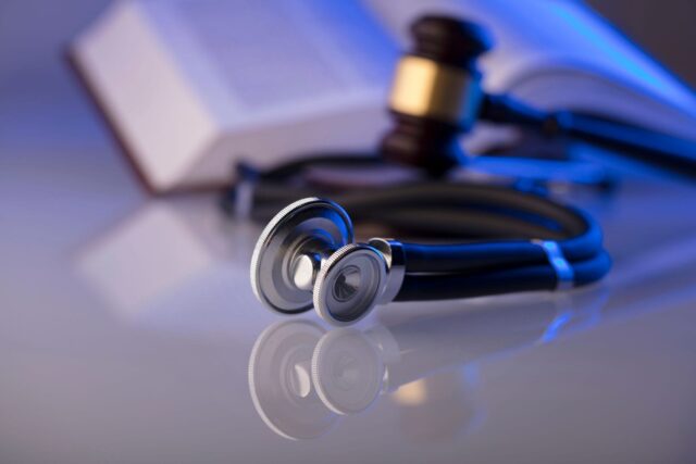 A stethoscope lying on a reflective surface with a blurred open book and a gavel in the background, illuminated by blue lighting.