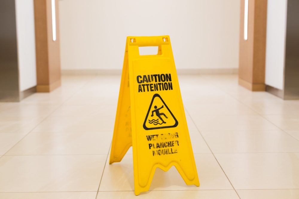 A yellow caution sign stands on a tiled floor in a hallway, warning of wet surfaces. Featuring a graphic of a person slipping and the text 
