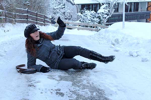 A person wearing winter clothes and a hat is slipping on an icy path, with an expression of surprise. In the background, there's snow-covered ground and a house surrounded by trees.